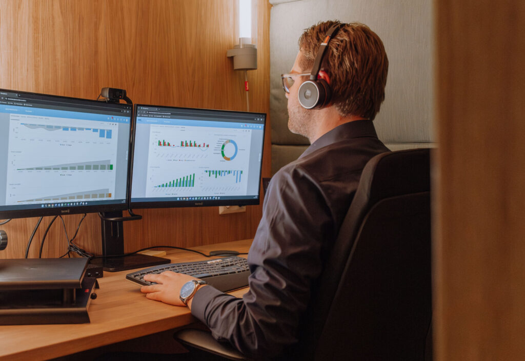 Man met koptelefoon werkt geconcentreerd aan bureau met twee schermen vol zakelijke grafieken en data-analyse, in een akoestische werkruimte.