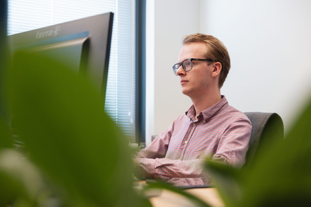 Medewerker met bril werkt geconcentreerd achter computerscherm op kantoor, gezien door groene planten op de voorgrond.