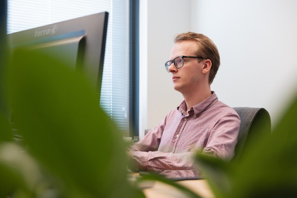 Medewerker met bril werkt geconcentreerd achter computerscherm op kantoor, gezien door groene planten op de voorgrond.