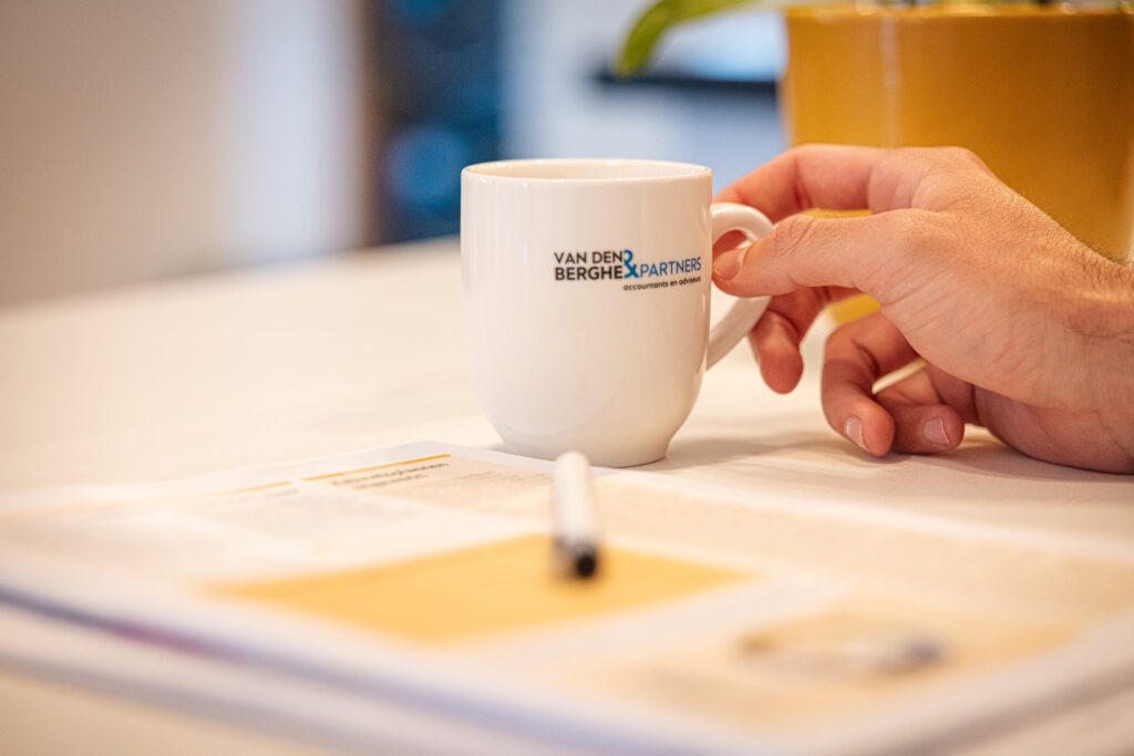 Hand pakt een koffiekop van Van den Berghe & Partners op tafel, met onscherpe documenten en pen op de voorgrond.