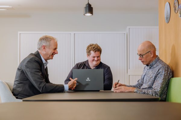 Drie mannelijke medewerkers in overleg rond een tafel, één typt op een laptop terwijl een ander aantekeningen maakt en de derde lacht mee.