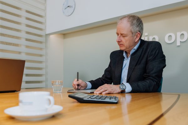 Man in pak maakt aantekeningen aan vergadertafel met rekenmachine, laptop en kop koffie, in een strak ingerichte kantoorruimte.
