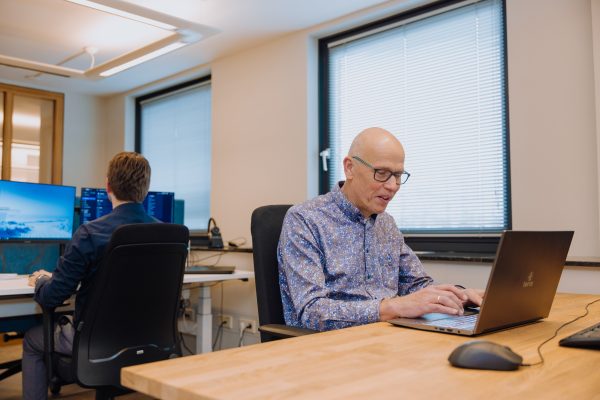 Twee mannen aan het werk op kantoor; één typt op een laptop, de ander werkt achter een scherm met meerdere monitoren.
