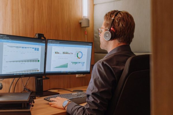 Man met koptelefoon werkt geconcentreerd aan bureau met twee schermen vol zakelijke grafieken en data-analyse, in een akoestische werkruimte.
