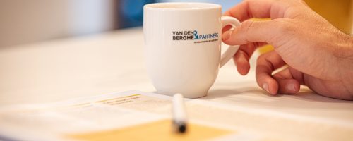 Hand pakt een koffiekop van Van den Berghe & Partners op tafel, met onscherpe documenten en pen op de voorgrond.