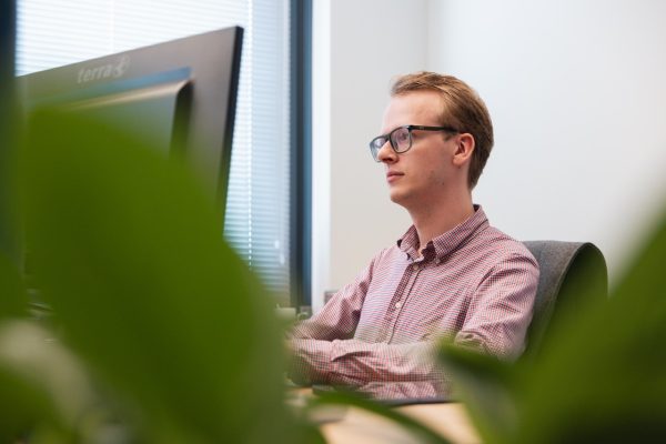 Medewerker met bril werkt geconcentreerd achter computerscherm op kantoor, gezien door groene planten op de voorgrond.