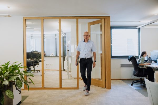 Man in lichte blouse en donkere broek loopt met een laptop onder de arm een moderne kantoorruimte uit, met glazen vergaderruimte op de achtergrond.